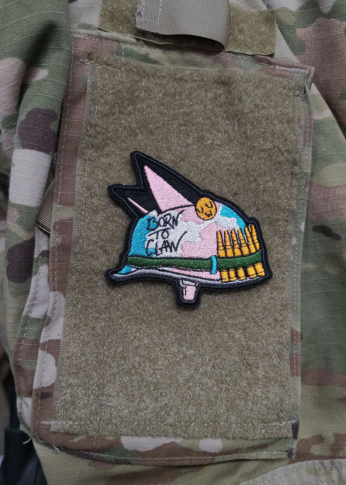A velcro patch in the shape of an Army helmet with cat ears. The camoflague is in the pastel colors of the trans flag. Written on the side is Born to Claw