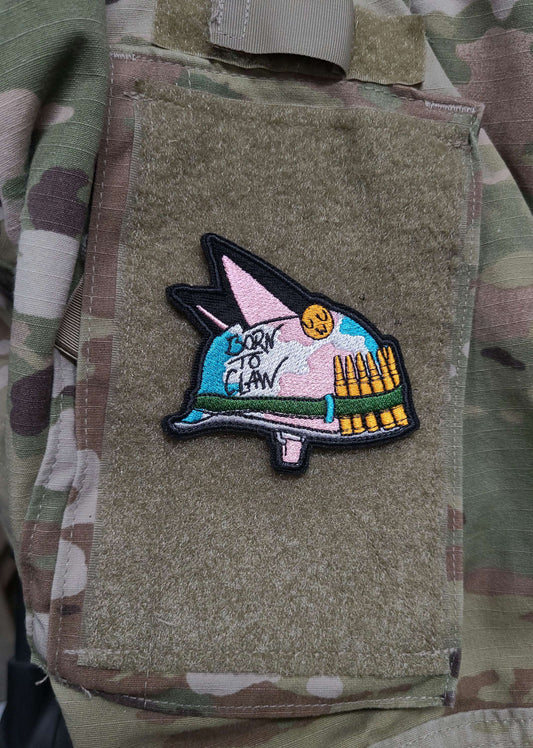 A velcro patch in the shape of an Army helmet with cat ears. The camoflague is in the pastel colors of the trans flag. Written on the side is Born to Claw