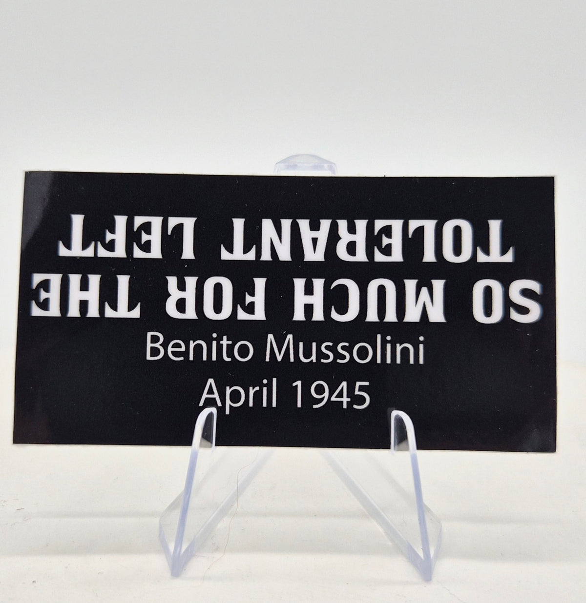 A black rectangular sign with white text reading 'SO MUCH FOR THE TOLERANT LEFT' upside down, followed by 'Benito Mussolini' and 'April 1945', displayed on a clear acrylic stand against a white background.